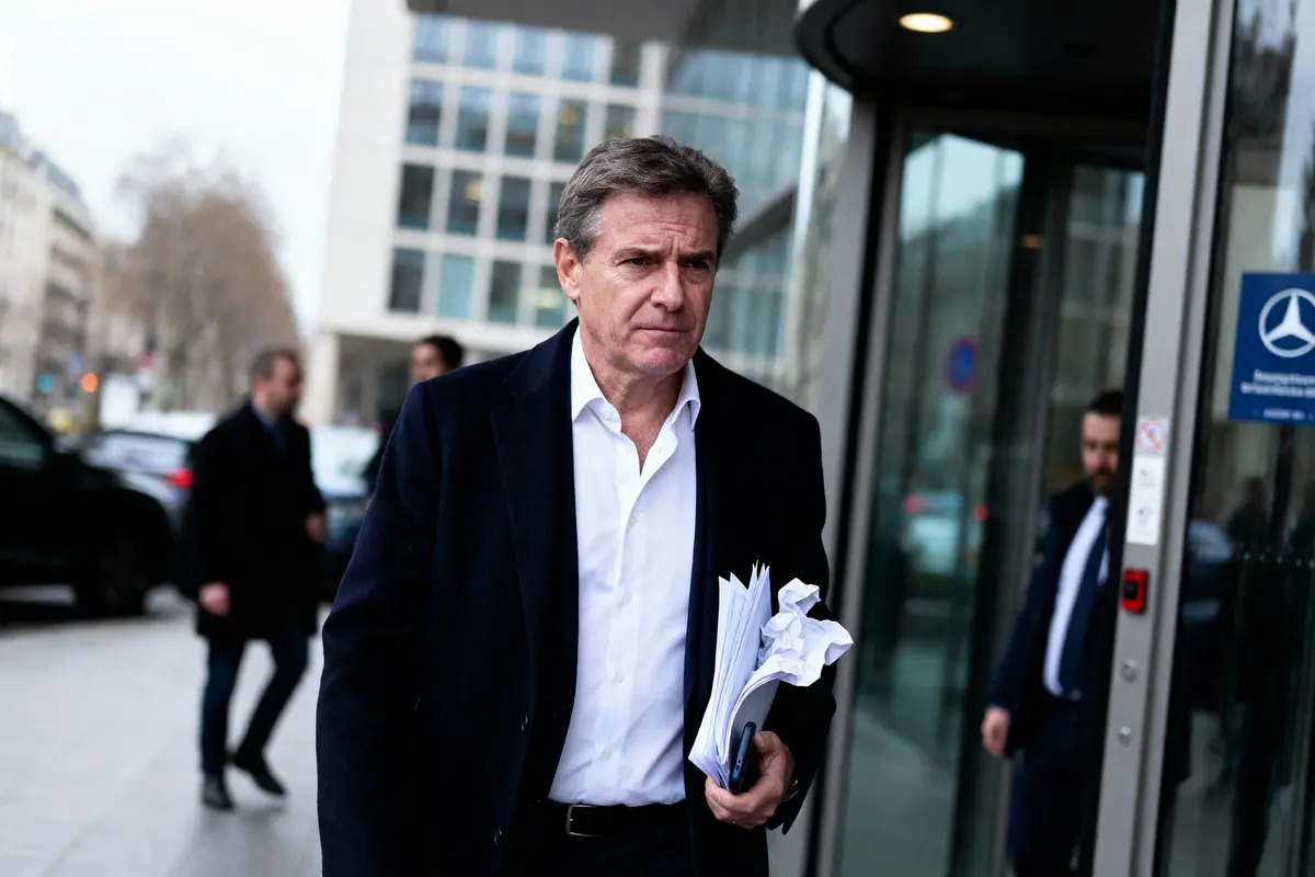 Un homme en costume, tenant des documents, sort d'un bâtiment moderne, entouré de voitures et passants flous.