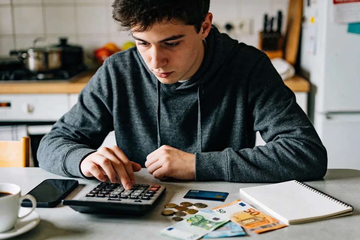 Jeune homme calculant budget sur calculatrice, avec billets, pièces, carte bancaire, cahier et smartphone sur table.