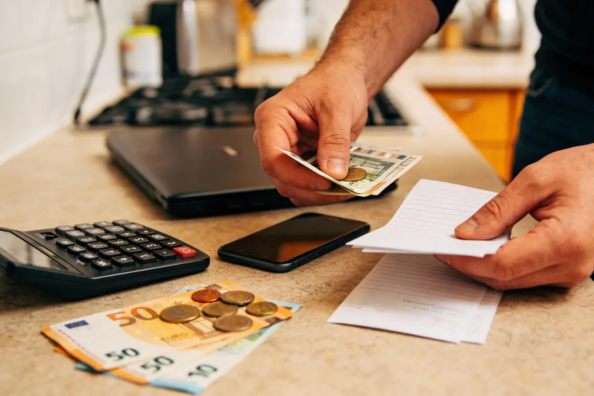 Une personne gère de l'argent, avec calculatrice, téléphone, billets et pièces sur une table de cuisine.
