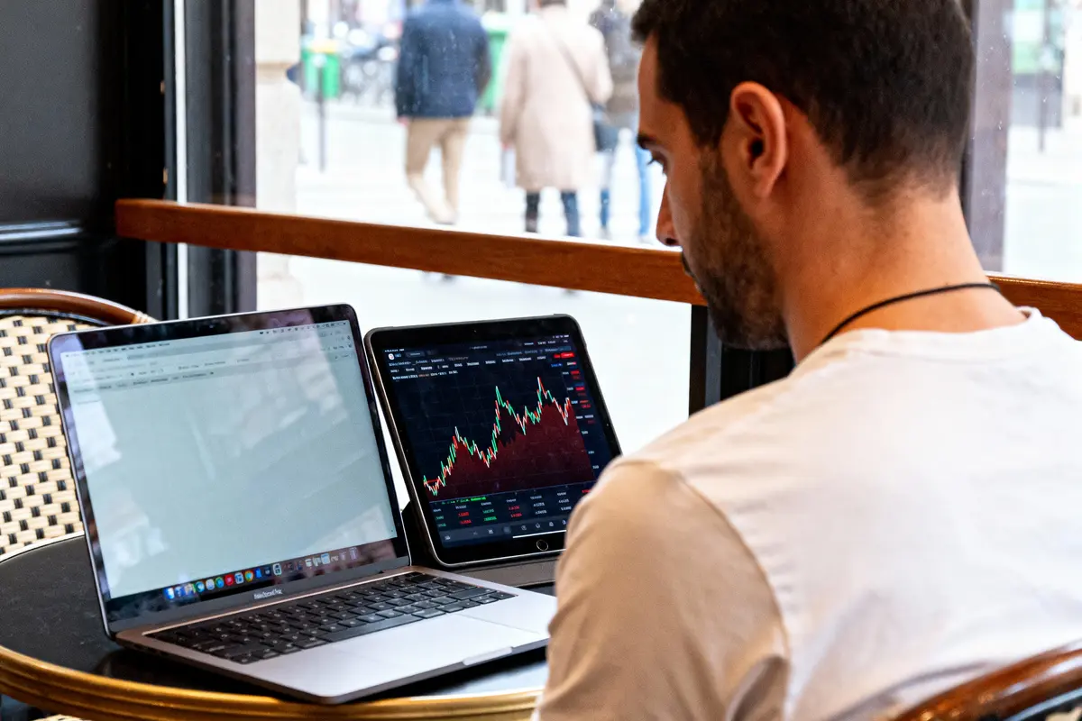 Homme utilisant deux ordinateurs portables dans un café, visualisant des graphiques financiers et du texte, vue de côté.
