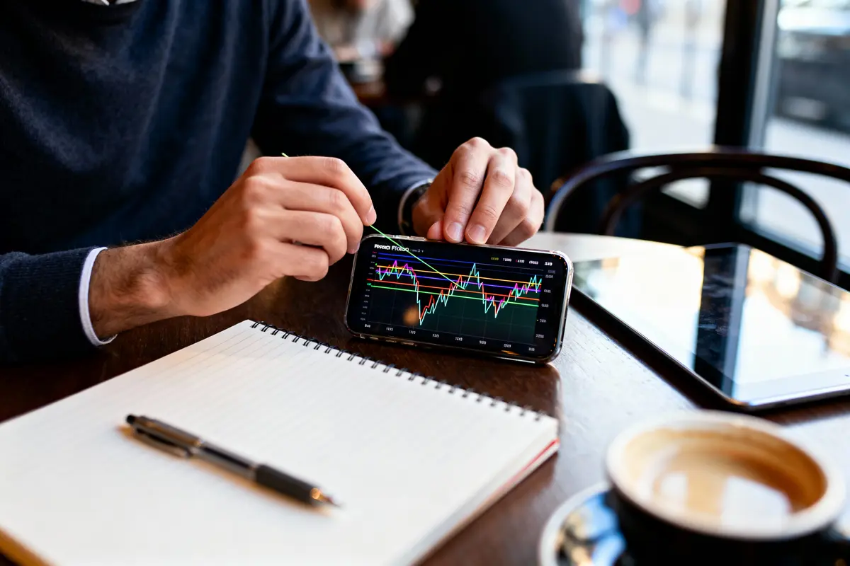 Personne analysant graphiques financiers sur smartphone, avec bloc-notes, stylo, tablette, tasse de café, dans un café.