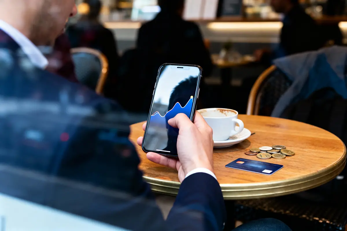 Personne tenant un smartphone affichant un graphique, assis à une table de café avec une carte bancaire.