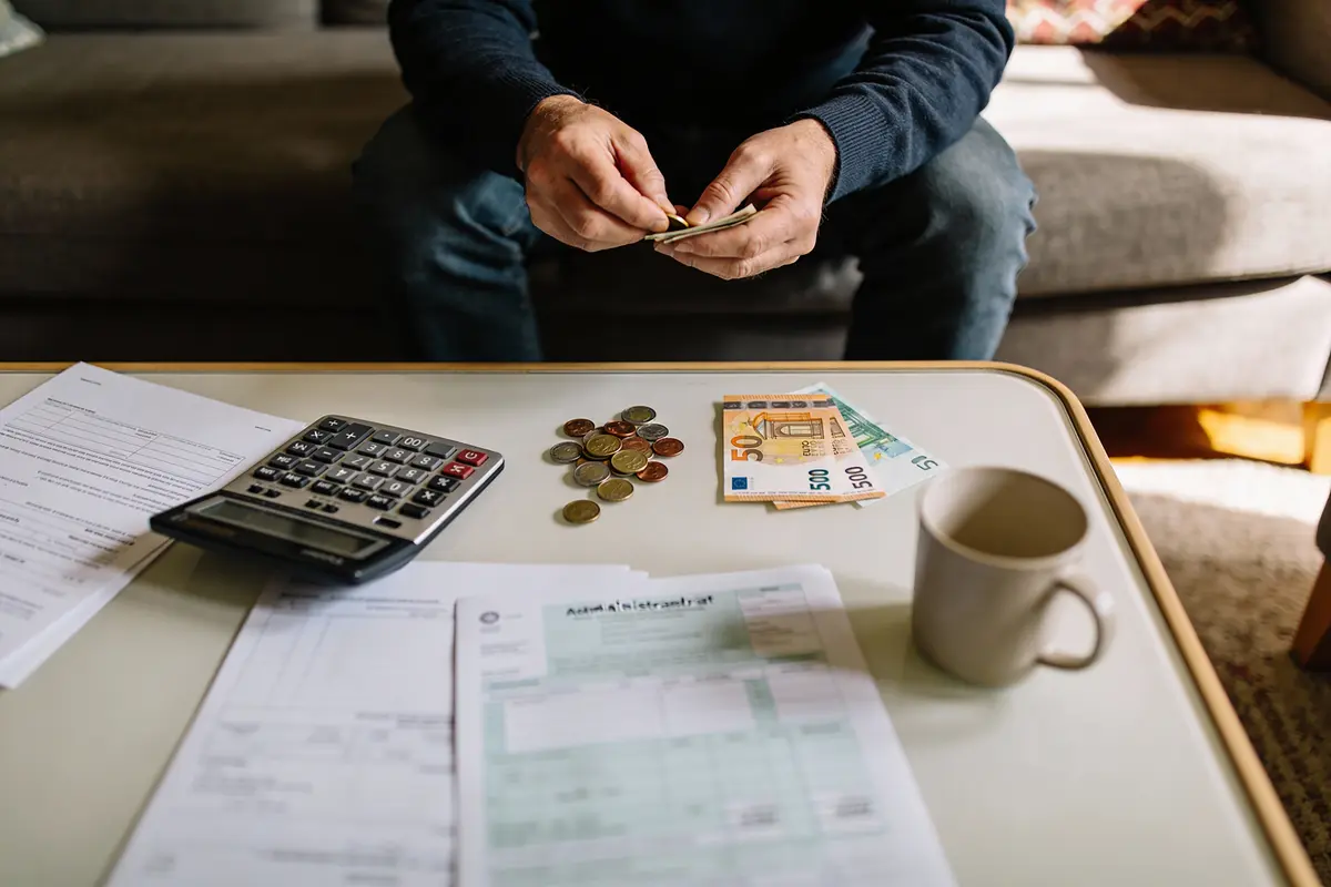 Personne calculant des finances à la table avec des billets, pièces, documents et une tasse.