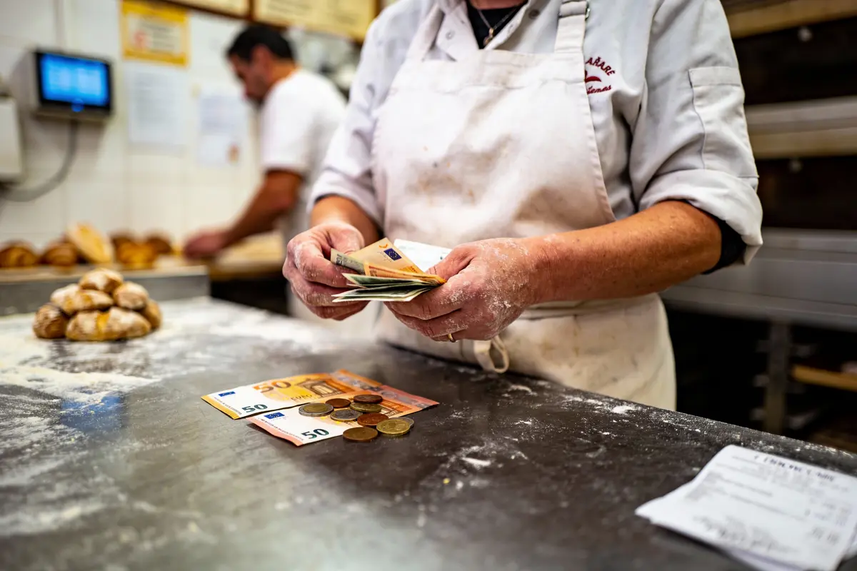 Boulangère comptant des billets et pièces sur un comptoir de boulangerie, farine et pain visibles.