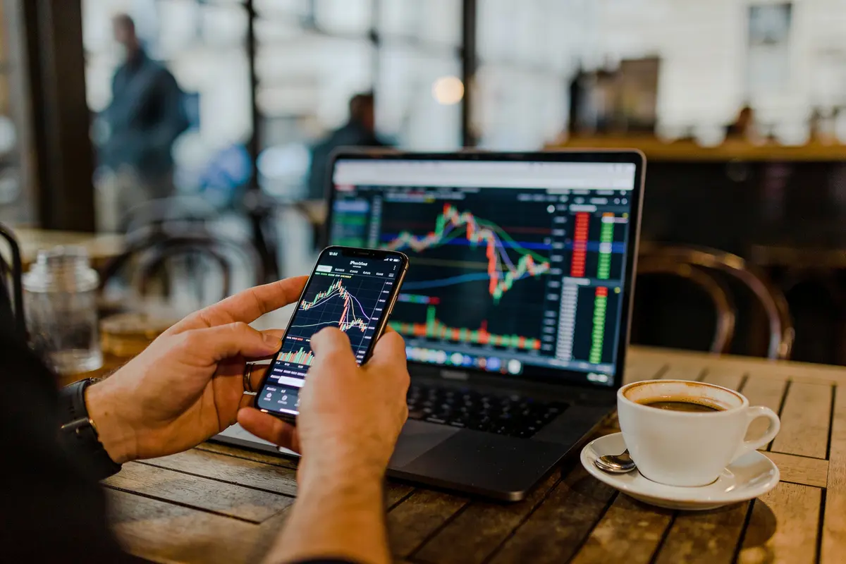 Personne analysant graphiques financiers sur smartphone et ordinateur portable dans un café. Tasse de café visible.