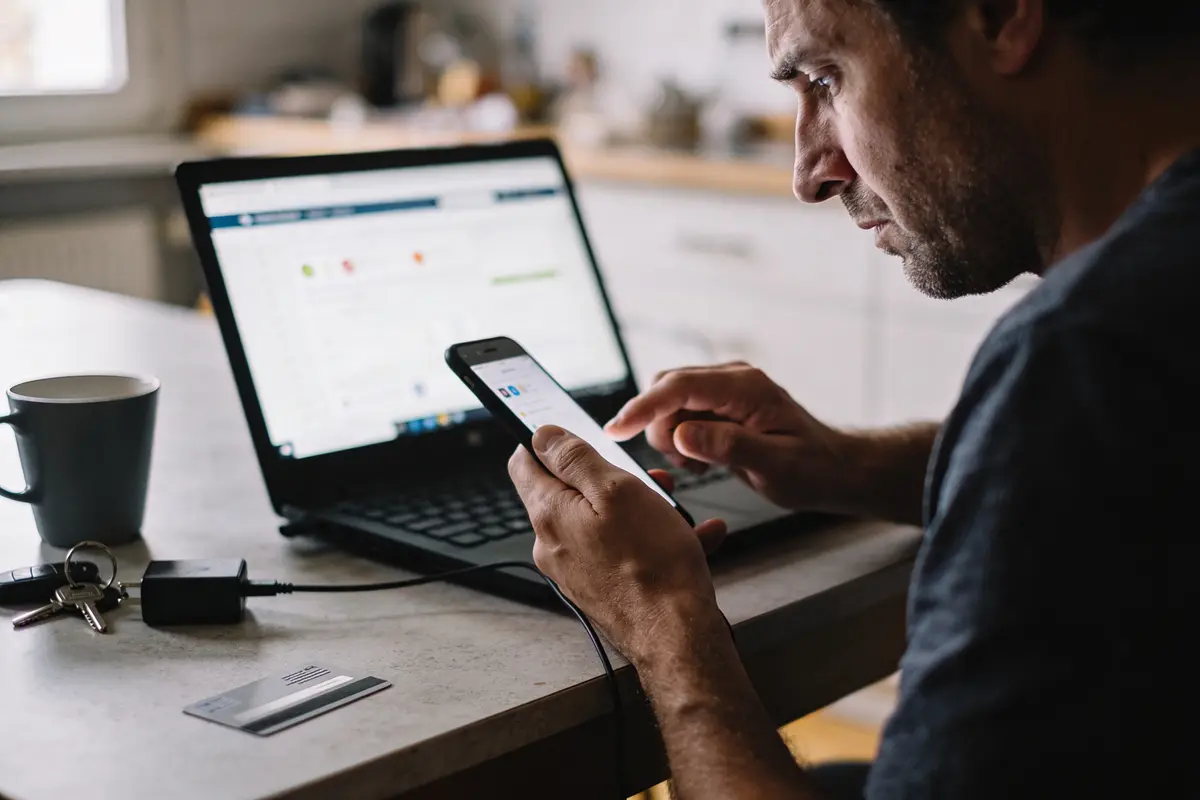 Homme utilisant smartphone et ordinateur portable sur table, tasse à proximité.