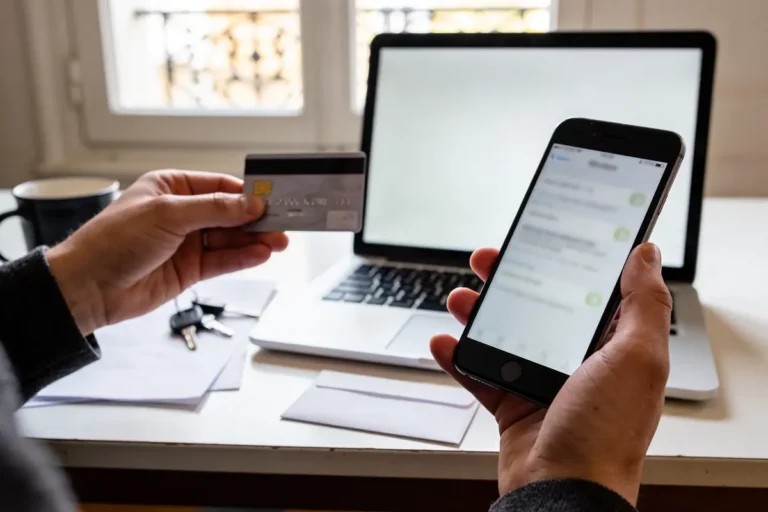 Personne tenant une carte bancaire et un smartphone devant un ordinateur portable sur un bureau.