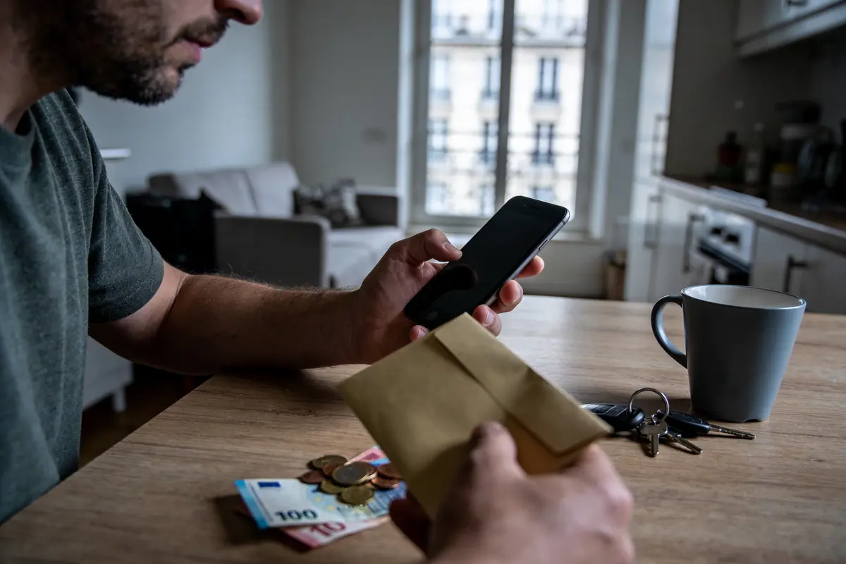 Homme utilisant un téléphone près d'enveloppe, argent, clés et tasse, dans un salon lumineux.