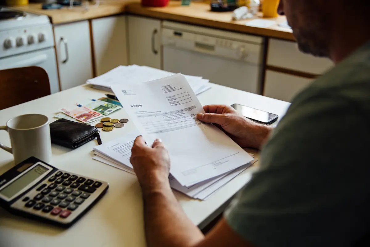 Personne lisant des factures à table, avec calculatrice, billets, pièces de monnaie, téléphone et tasse.