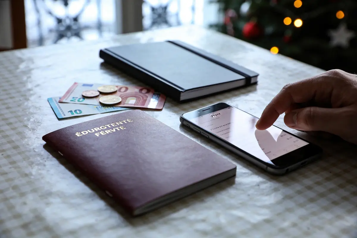 Passeport, argent, téléphone sur table, cahier noir, sapin de Noël en arrière-plan, main utilisant smartphone.