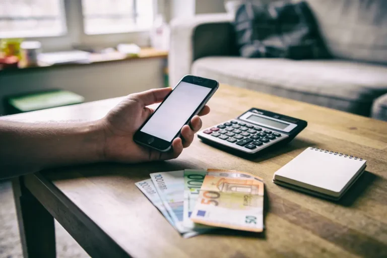 Main tenant smartphone près d'argent, calculatrice et carnet sur table en bois, intérieur lumineux.
