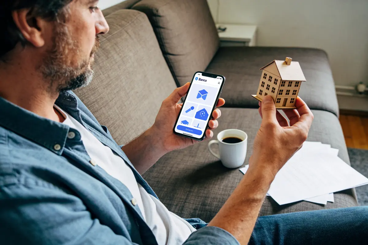 Homme tient une maison miniature et smartphone avec application bancaire, assis sur un canapé avec café.