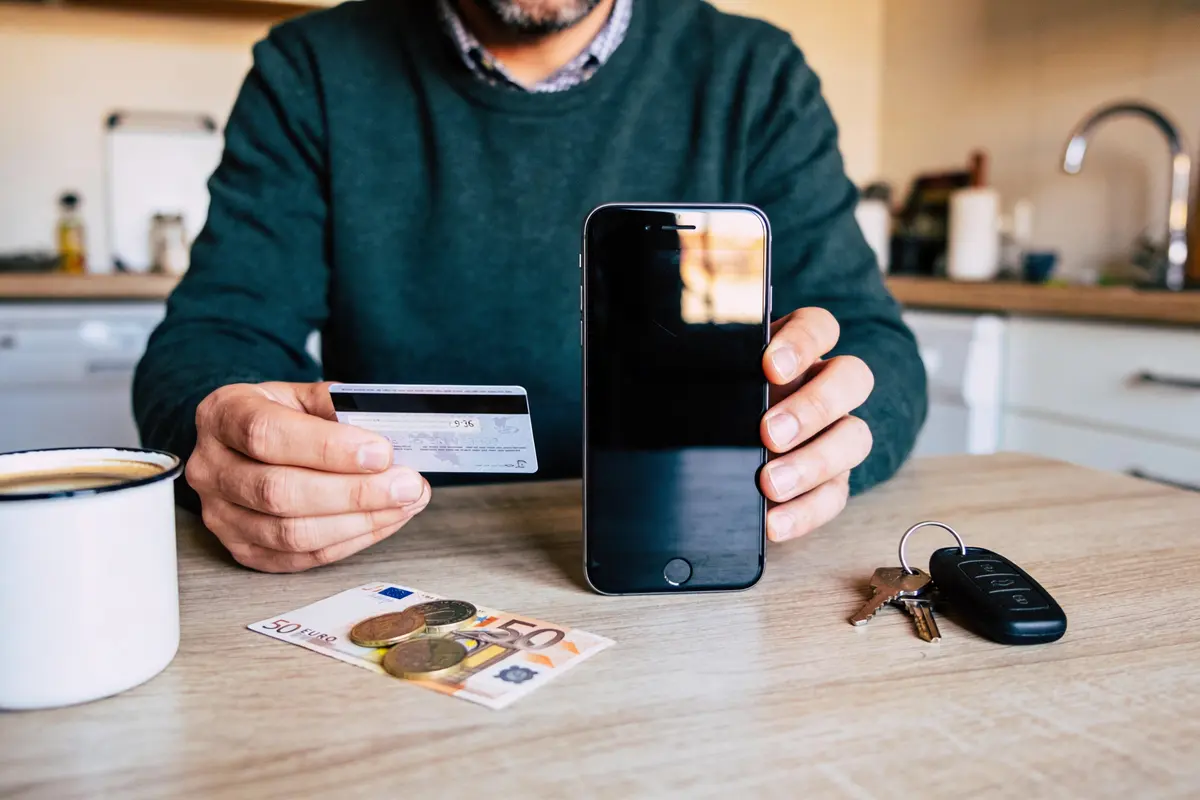 Homme tenant smartphone et carte bancaire, argent sur table, tasse de café et clés visibles.