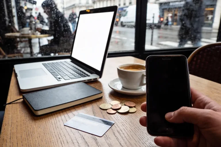 Ordinateur, smartphone, carnet, café, pièces, carte bancaire sur table en bois, fenêtre mouillée, rue animée en arrière-plan.