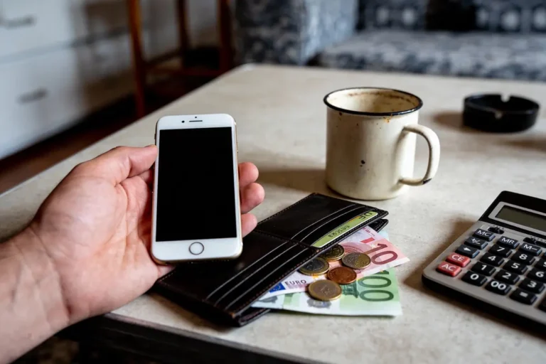 Main tenant smartphone, portefeuille avec euros, tasse émaillée, calculatrice sur table blanche.