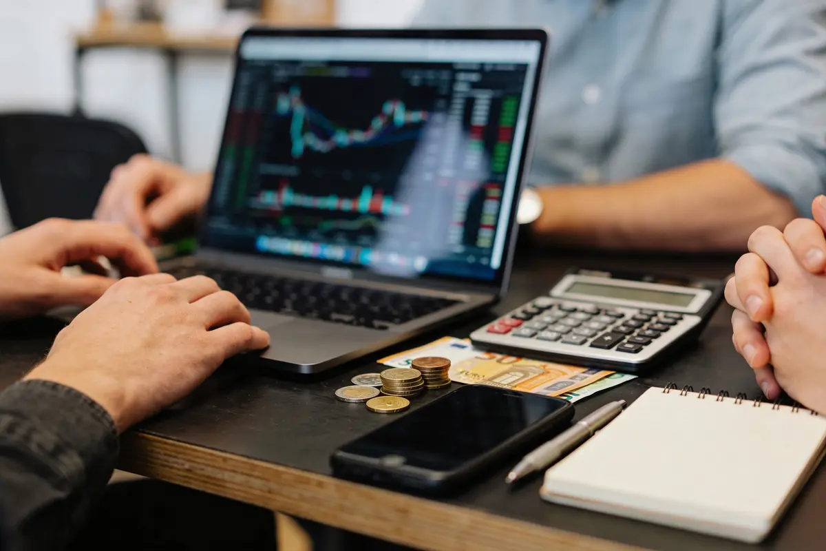 Personnes travaillant sur ordinateur portable avec graphique, calculatrice, pièces de monnaie, billets, téléphone et cahier sur table.