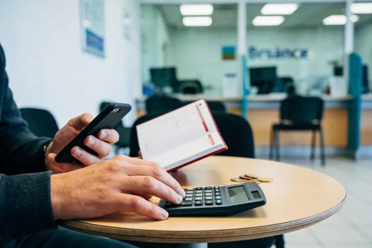 Personne utilisant calculatrice et téléphone avec un carnet, pièces sur table, dans un espace de bureau.