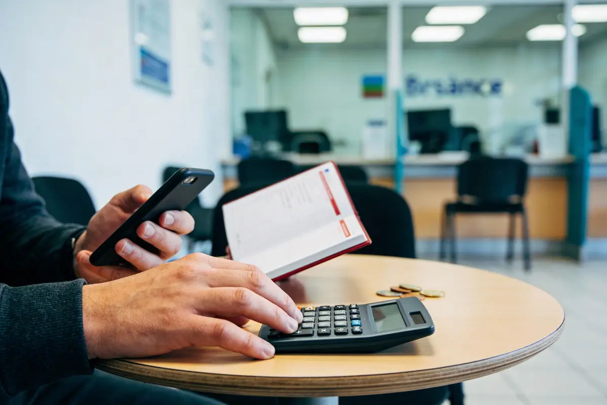 Personne utilisant calculatrice et téléphone avec un carnet, pièces sur table, dans un espace de bureau.