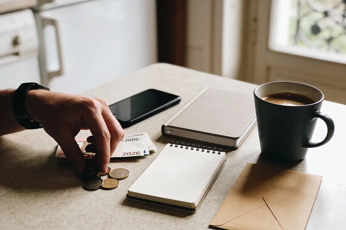 Main, pièces de monnaie, billets, téléphone, carnet, tasse de café sur table lumineuse. Atmosphère décontractée et organisée.