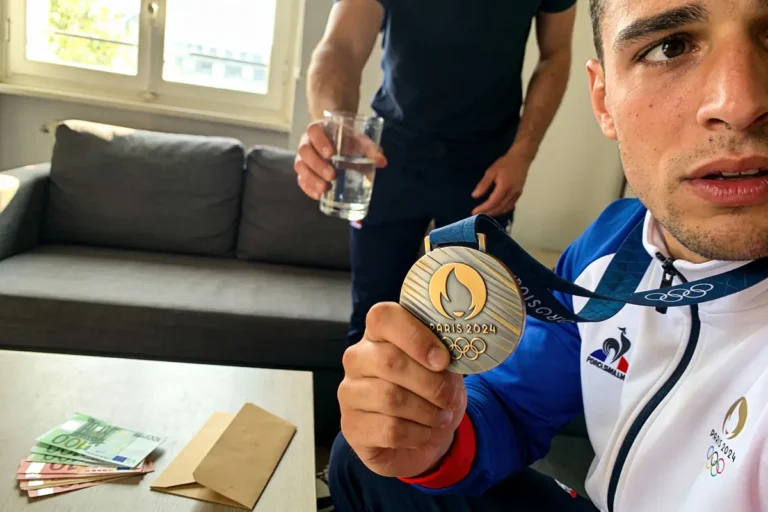 Homme tenant médaille Paris 2024, verre d'eau, billets et enveloppe sur la table.