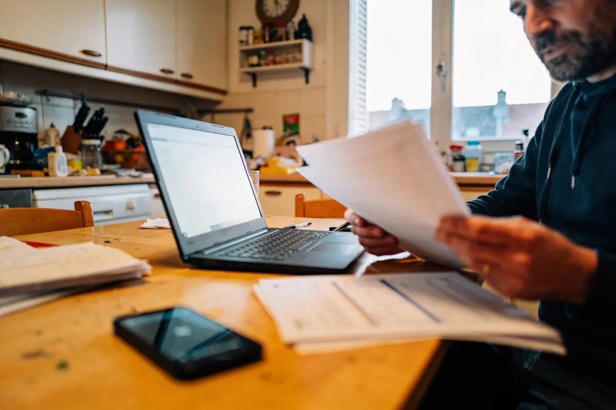 Homme travaillant à domicile, tenant des documents, ordinateur portable sur table, intérieur de cuisine en arrière-plan.