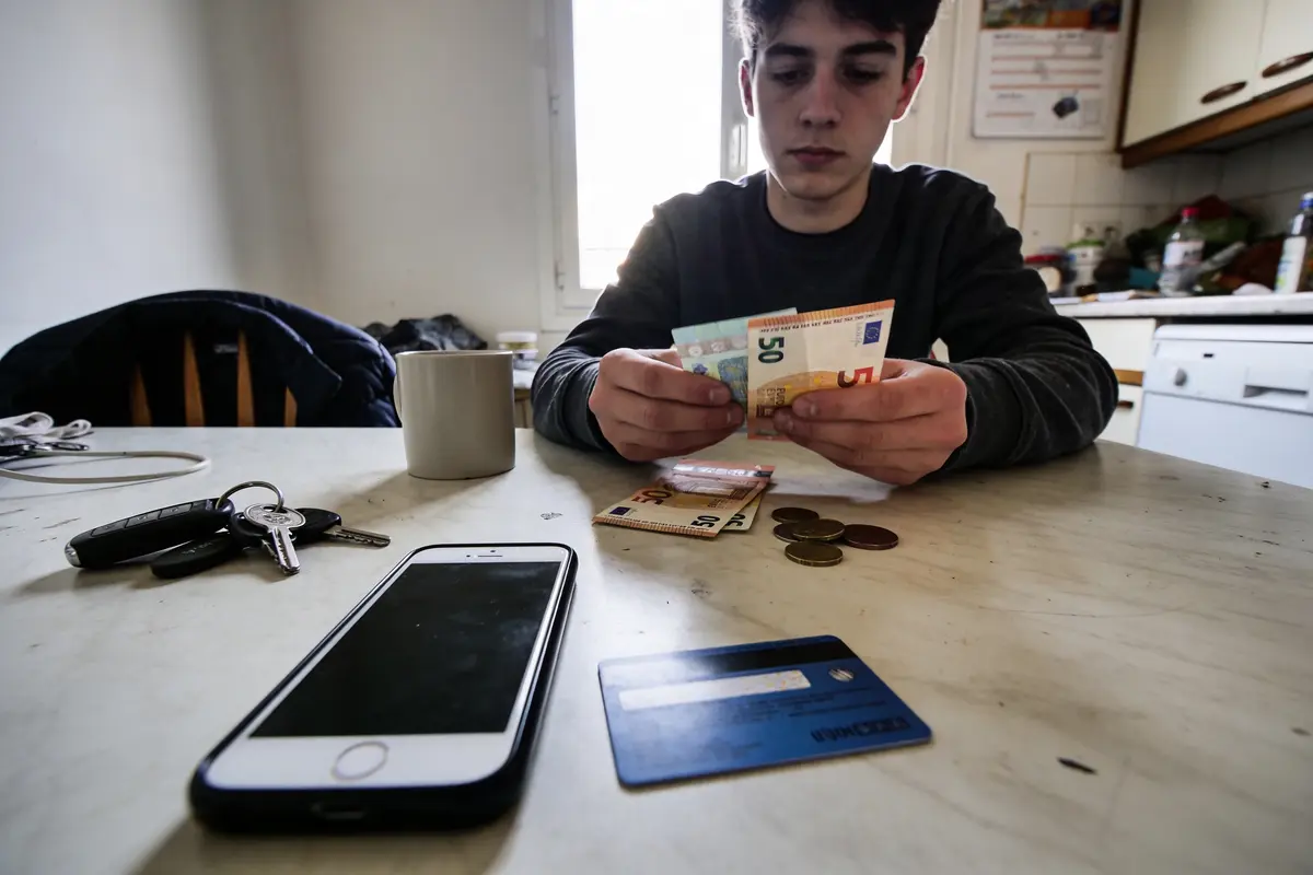 Un jeune homme compte des billets sur une table avec téléphone, clés, carte bancaire et tasse.