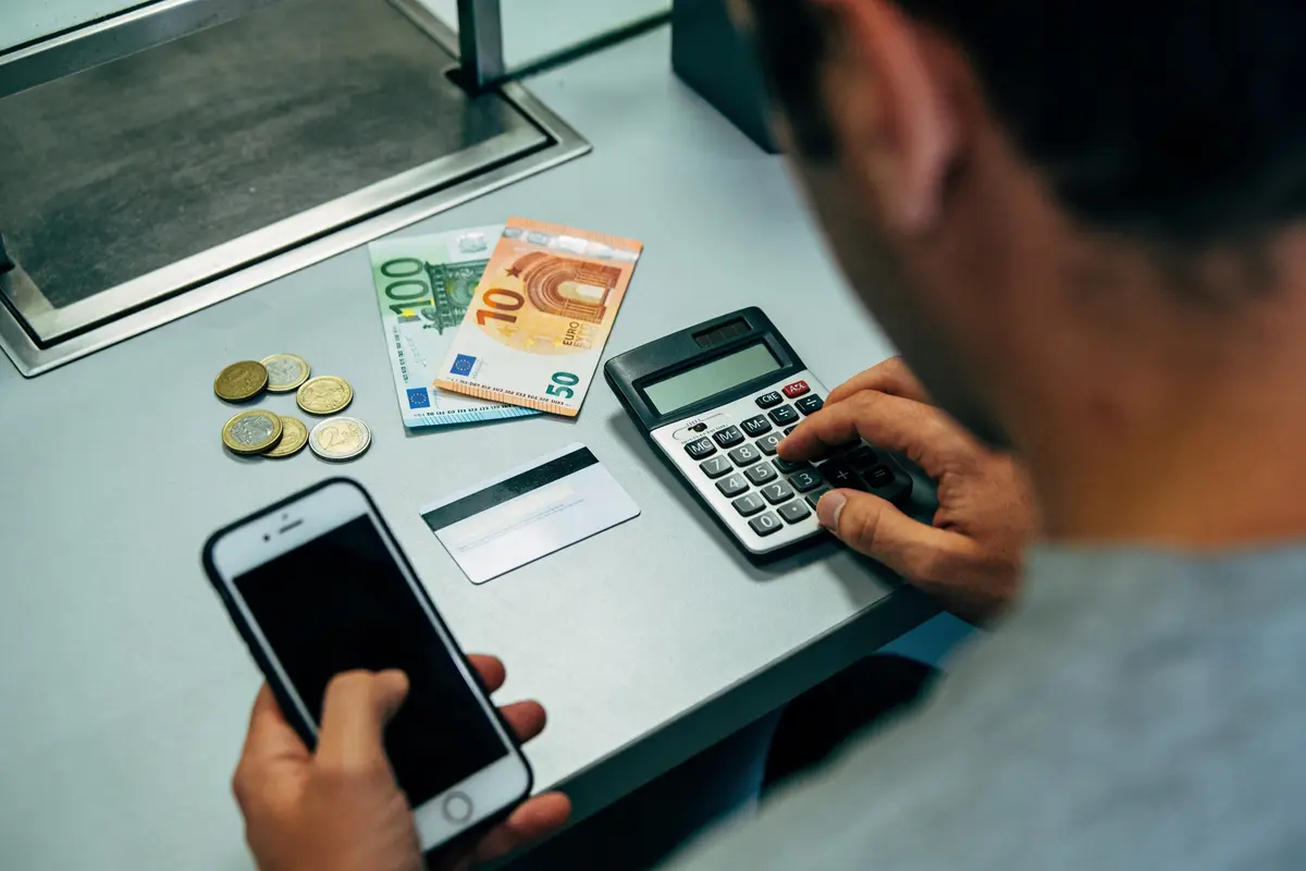 Personne utilisant calculatrice et smartphone, avec argent en euros et carte bancaire sur table grise.