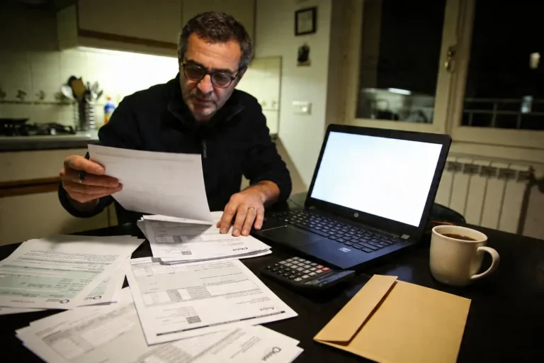 Un homme lit des documents, ordinateur portable allumé, avec une calculatrice et un café à côté.