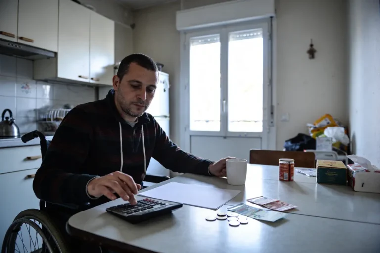 Un homme en fauteuil roulant utilise une calculatrice à table, entouré de billets, pièces et tasses.