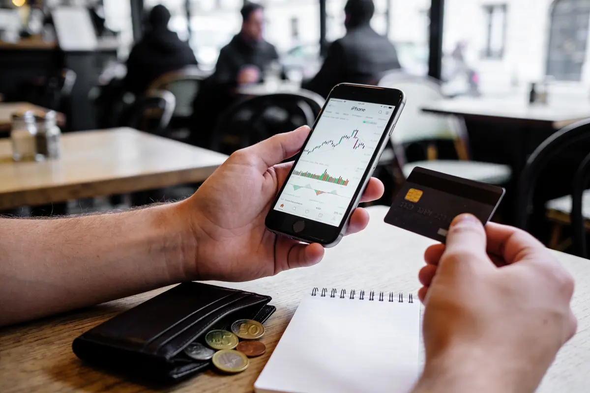 Personne tient smartphone avec graphique et carte bancaire, café en arrière-plan, carnet et pièces sur la table.