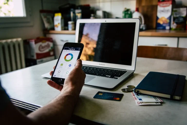 Personne utilisant smartphone avec graphique, près d'un ordinateur portable, carte bancaire, pièces et carnet sur table.