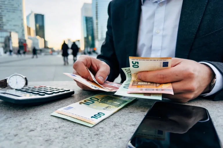 Personne comptant des billets en euros, avec calculatrice, montre et téléphone sur une table extérieure en ville.