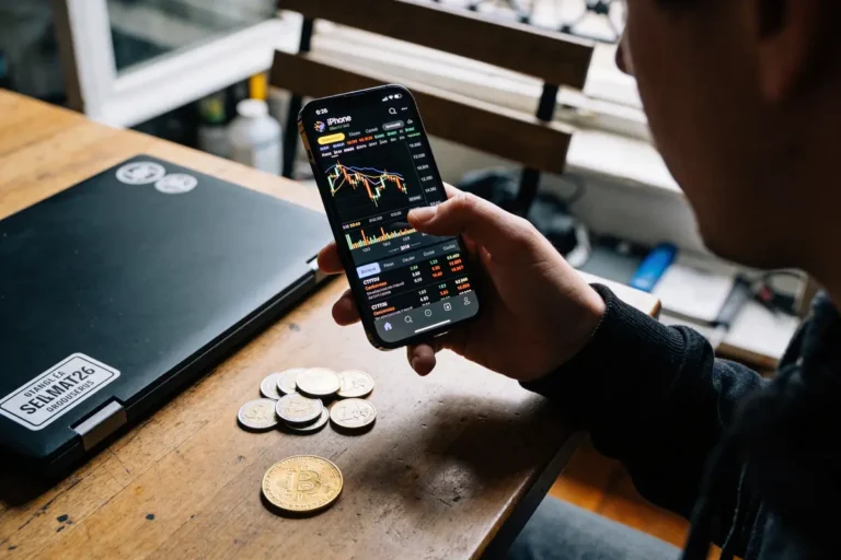 Personne consultant des graphiques boursiers sur smartphone, pièces de monnaie et ordinateur portable sur table en bois.