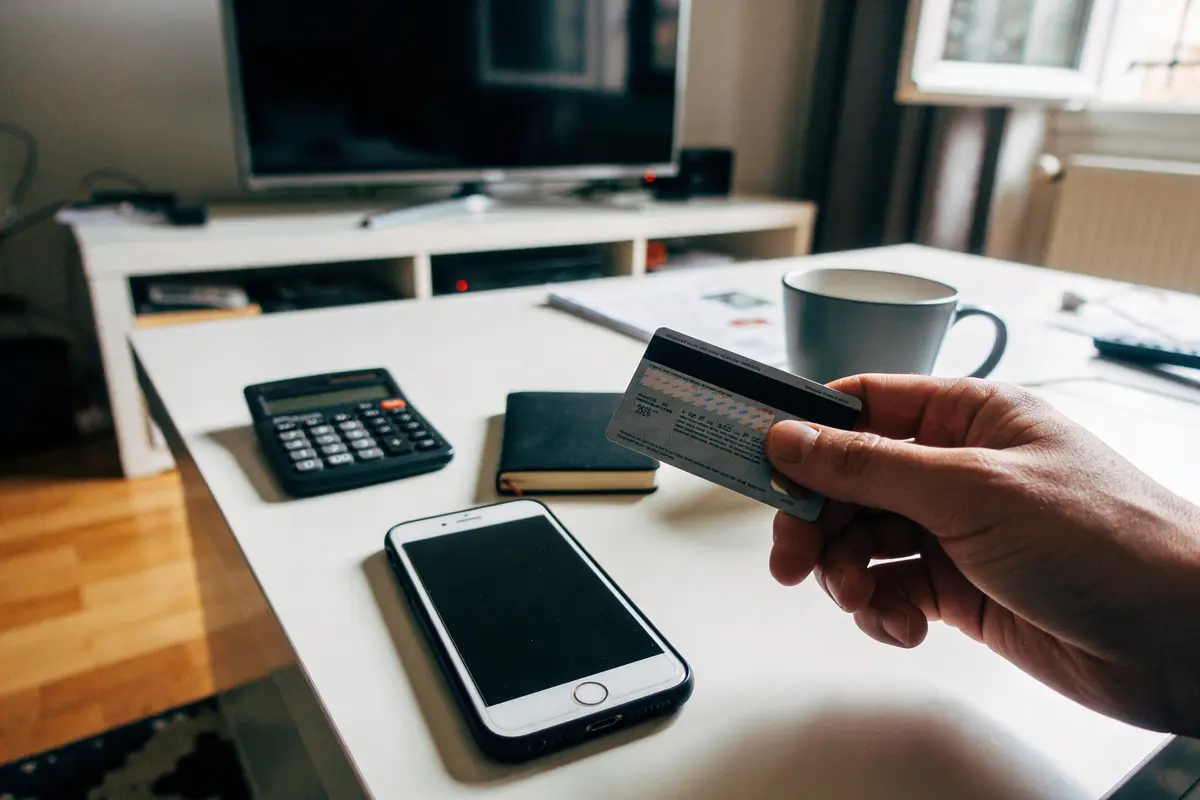 Main tenant une carte de crédit, avec smartphone, calculatrice, carnet et tasse sur table.