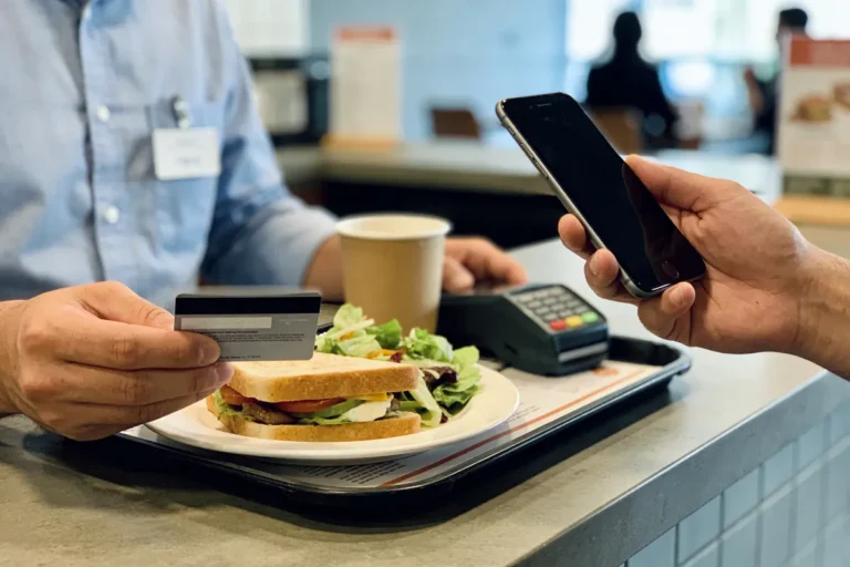 Paiement mobile sans contact dans un café : personne tenant smartphone, carte bancaire, sandwich et boisson.