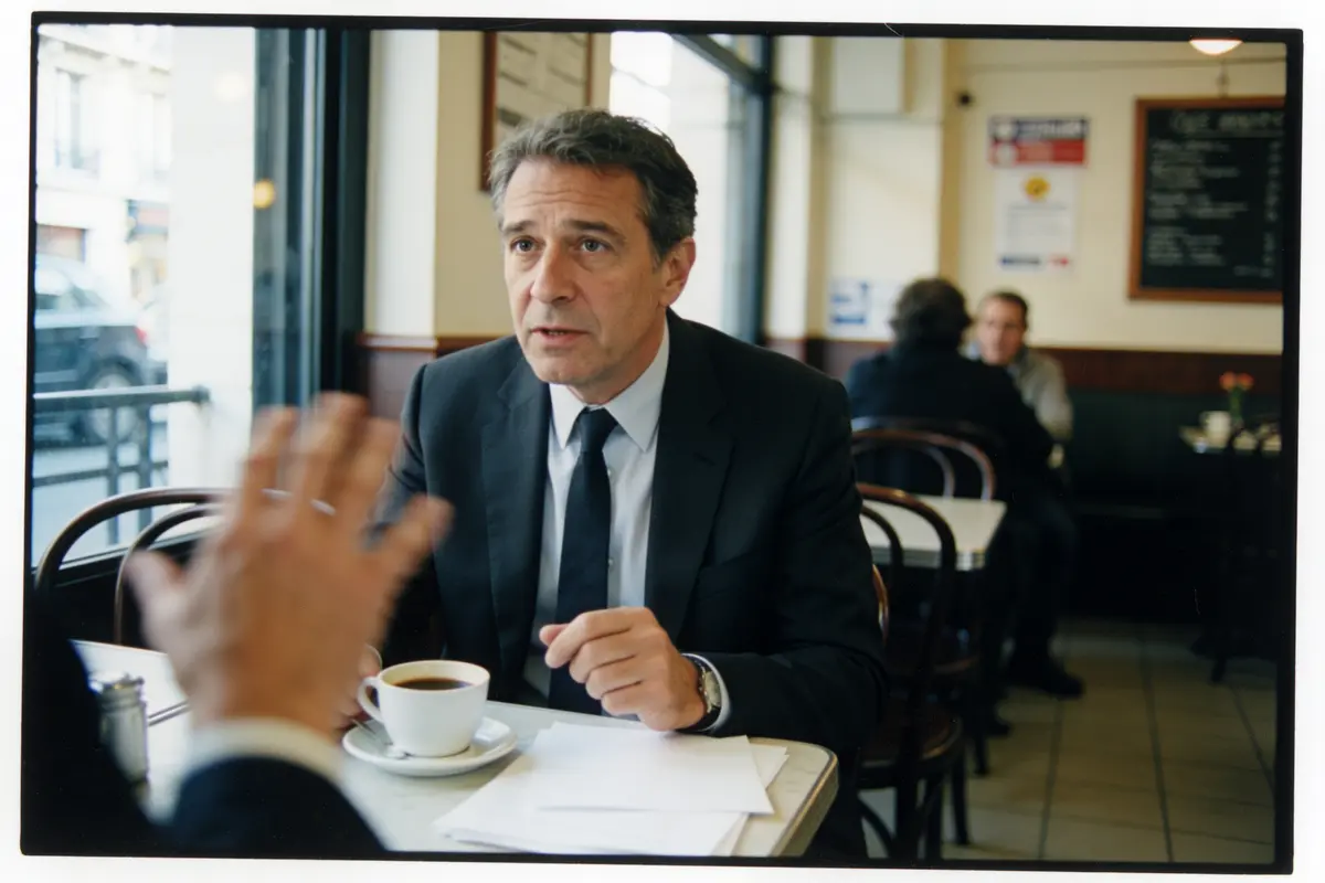 Un homme en costume boit un café dans un café, discutant avec quelqu'un en face.