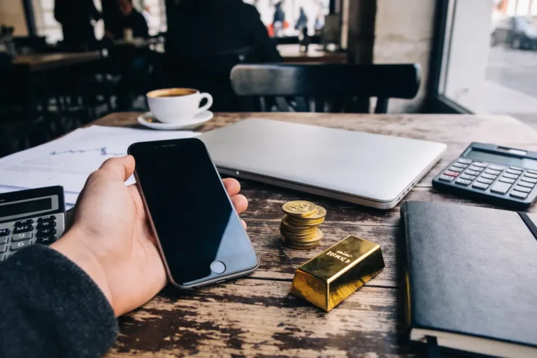 Bureau avec smartphone, ordinateur, lingot d'or, pièces empilées, calculatrice et cahier sur une table en bois.