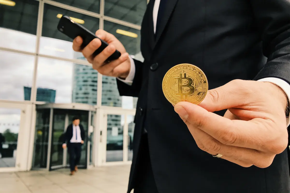 Homme en costume tenant un téléphone et un bitcoin devant un bâtiment moderne.