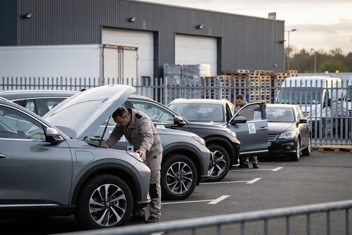 Personnes inspectant des voitures alignées sur un parking industriel, capots ouverts, bâtiment métallique en arrière-plan.