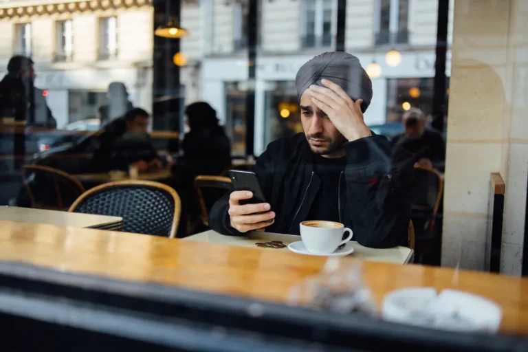 Homme pensif regarde son téléphone dans un café, tasse de café et pièces devant lui.