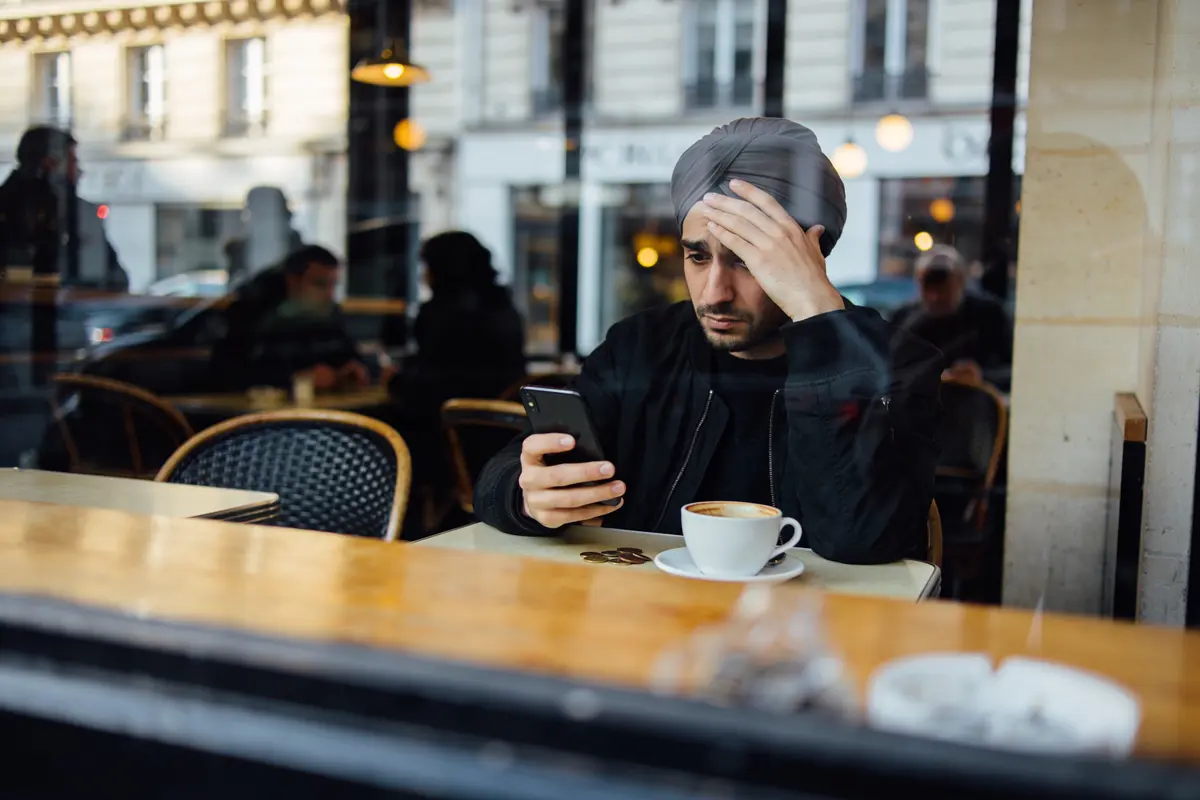 Homme pensif regarde son téléphone dans un café, tasse de café et pièces devant lui.