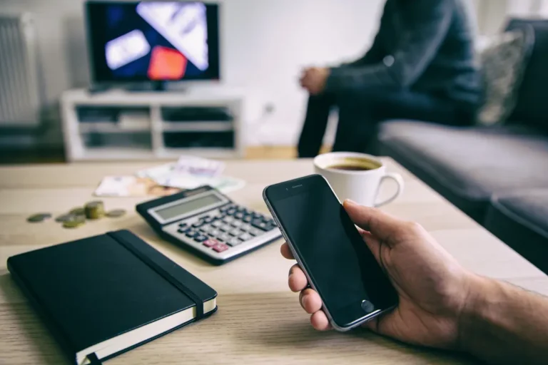 Personne tenant un smartphone, table avec calculatrice, carnet, tasse de café. Télévision floue en arrière-plan.