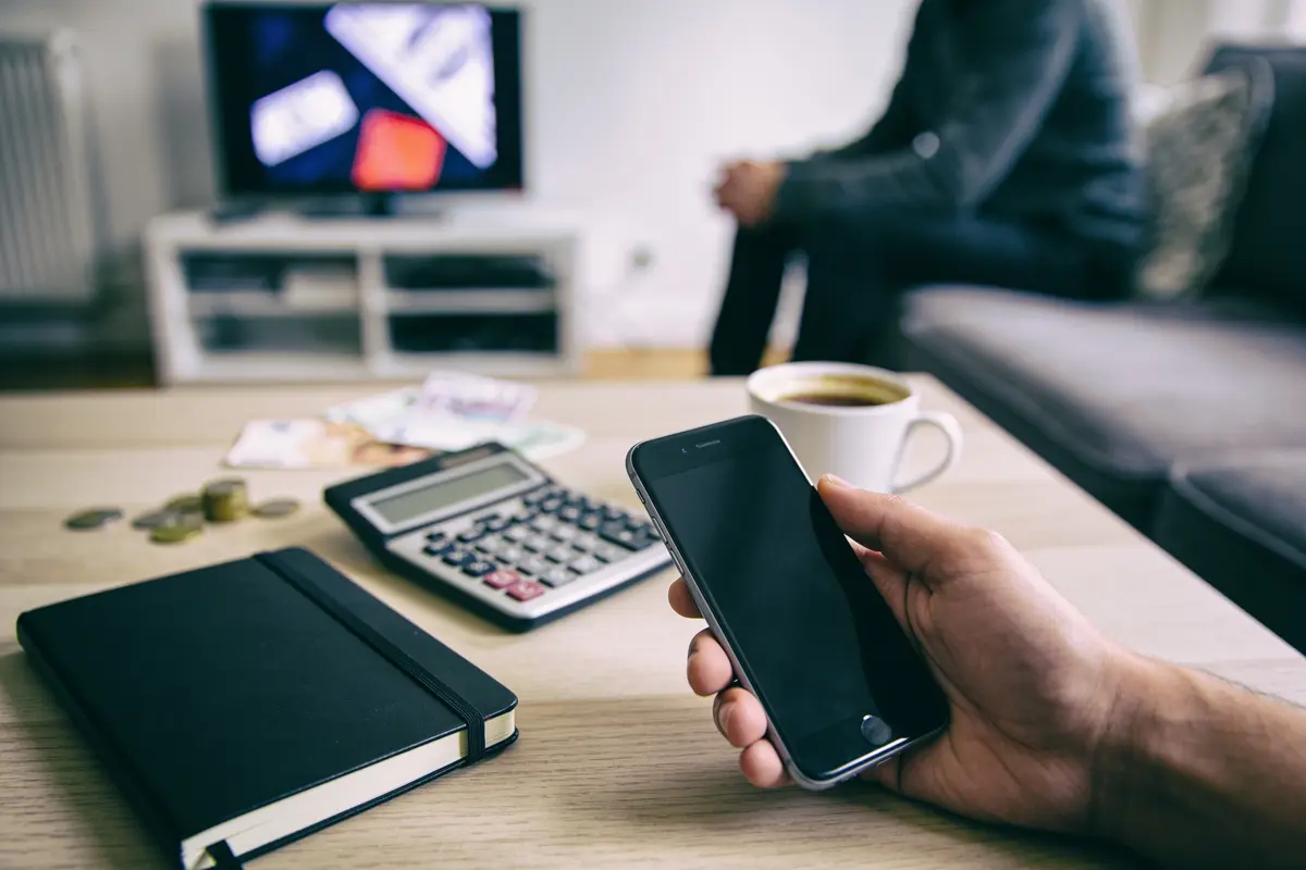 Personne tenant un smartphone, table avec calculatrice, carnet, tasse de café. Télévision floue en arrière-plan.
