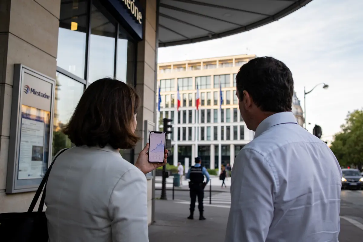 Deux personnes regardant un graphique sur un smartphone, près d'un bâtiment gouvernemental, policier au loin.