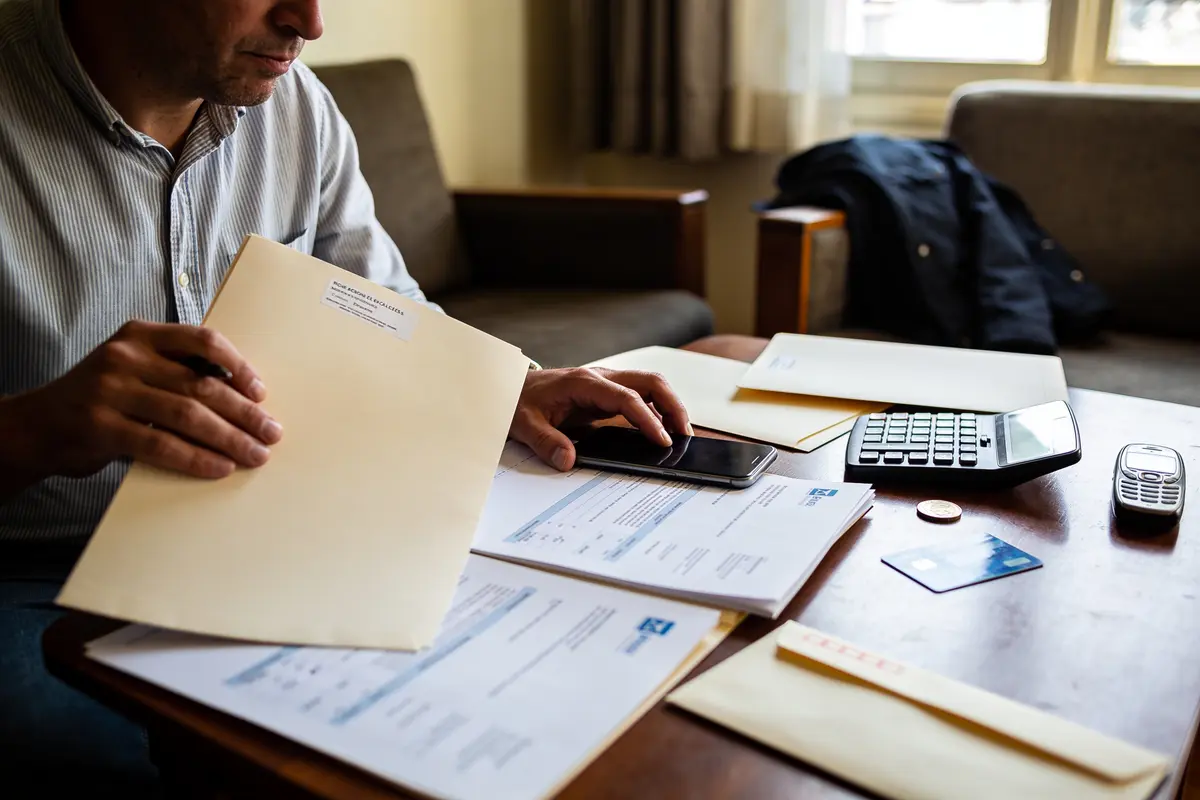 Homme gérant des documents administratifs sur une table avec calculatrice, téléphone et carte bancaire.