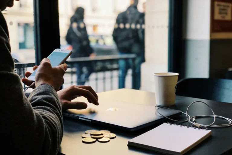 Personne utilisant smartphone et ordinateur portable dans un café, avec monnaie, carnet, et gobelet sur la table.