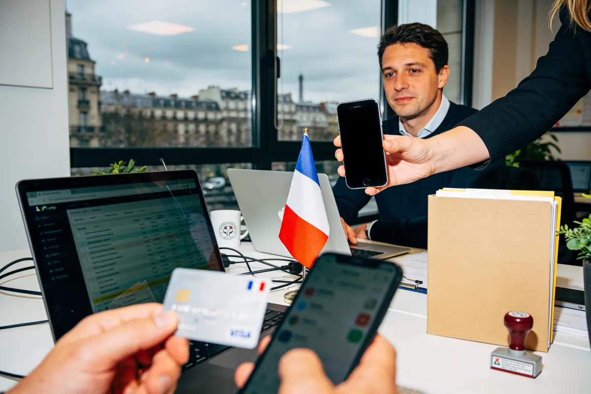 Bureau avec ordinateur, carte bancaire, drapeau français, smartphone, documents, homme regardant écran, fenêtres Paris en arrière-plan.