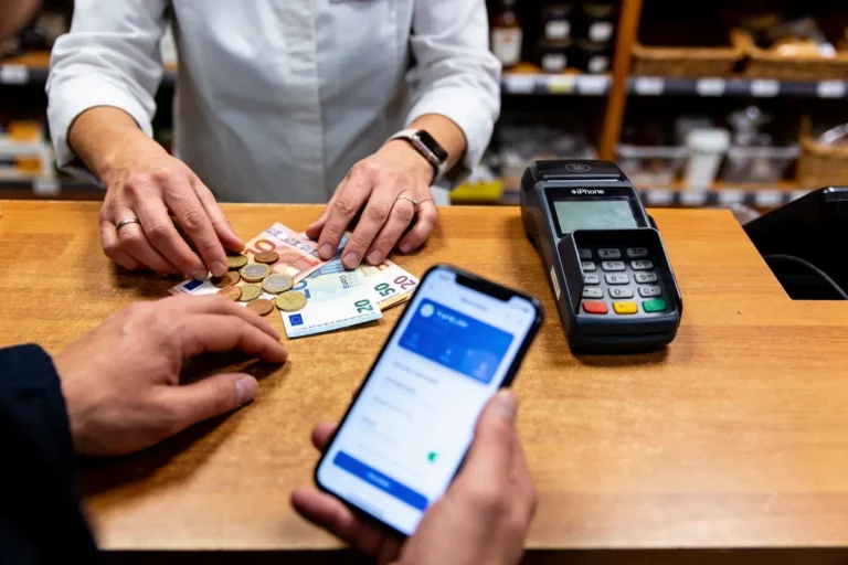 Personne effectuant un paiement avec smartphone, billets, et pièces devant un terminal de paiement en magasin.