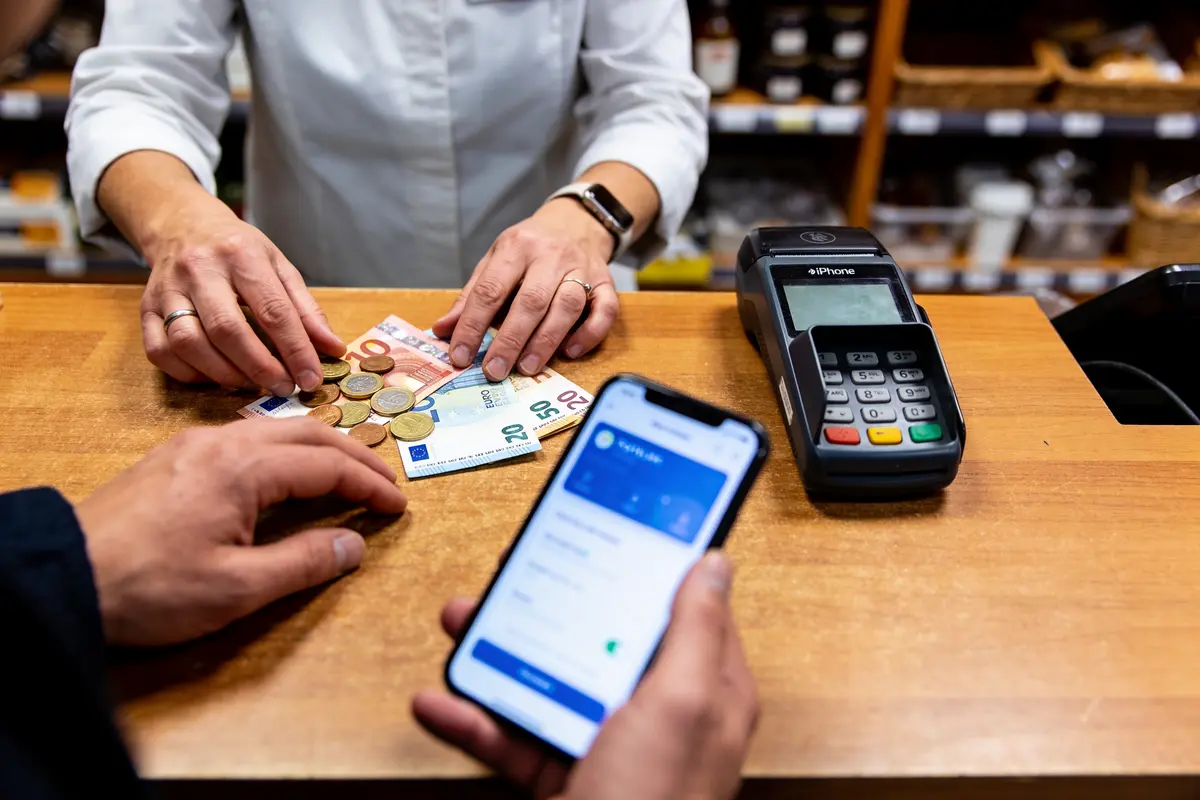 Personne effectuant un paiement avec smartphone, billets, et pièces devant un terminal de paiement en magasin.