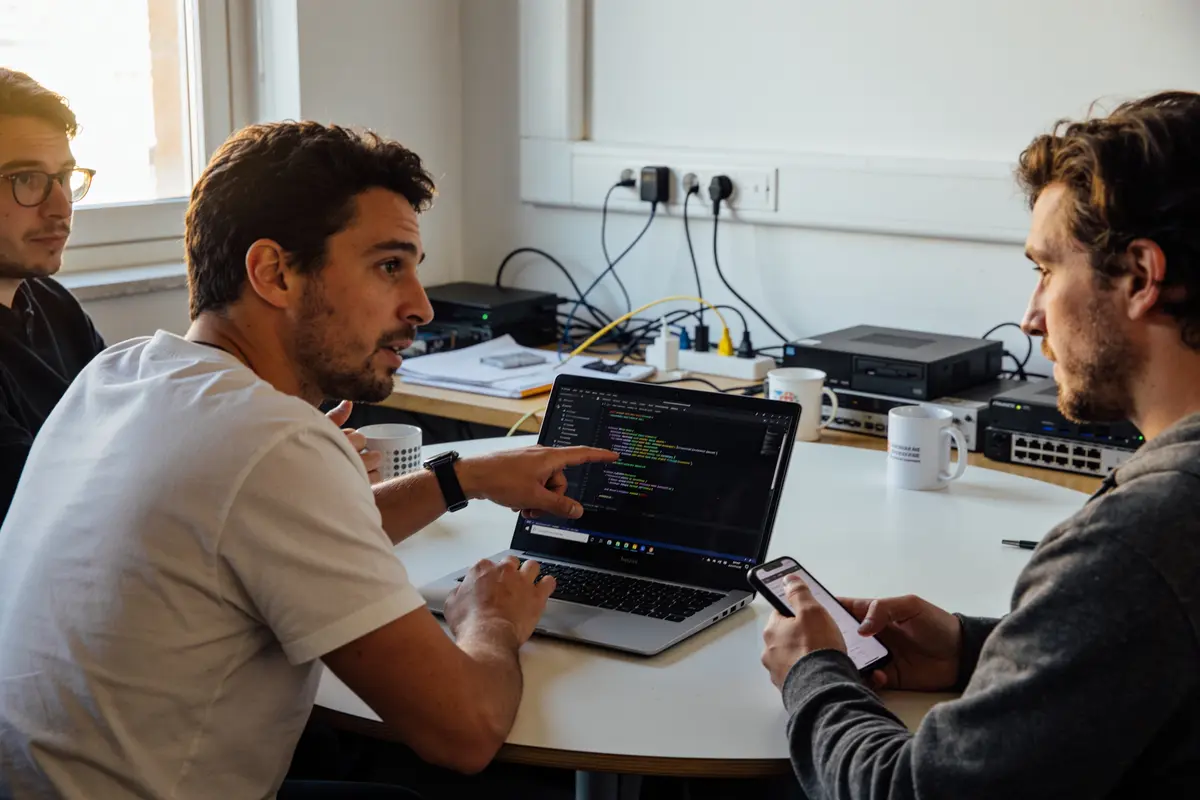 Trois hommes discutant de code informatique autour d'un ordinateur portable dans un bureau.